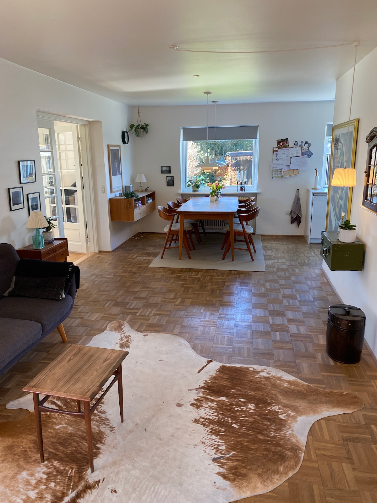 An open living area features a comfortable sofa, a small coffee table, and a decorative cowhide rug on the floor. A dining table with chairs is positioned near a window, allowing natural light to fill the space, showcasing a warm and functional atmosphere.