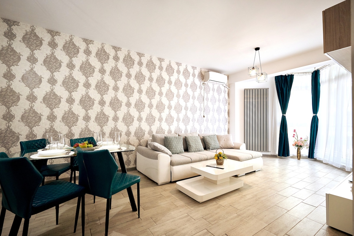 A spacious living area is furnished with a large sectional sofa and a modern coffee table. A dining table with blue chairs is positioned nearby. Floor-to-ceiling windows allow natural light to fill the room, while elegant wallpaper adds a touch of style.