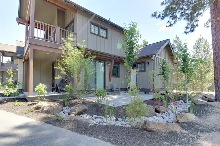 A modern two-story home is showcased, surrounded by landscaping that includes small rocks and various plants. A pathway leads from the driveway to the entrance, with a private seating area visible on the porch. The building features large windows that allow natural light to enter.