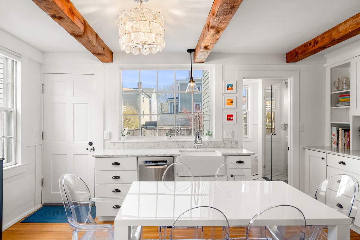 A modern kitchen is highlighted by a spacious white dining table and transparent chairs. Natural light enters through the window above the farmhouse sink, illuminating the marble countertops and contrasting with the wooden beams overhead. A doorway leads to a bright bathroom.