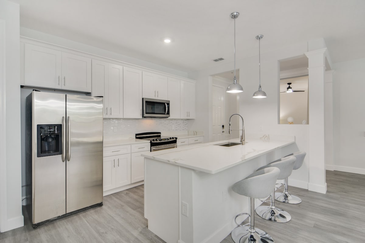 A modern kitchen is equipped with stainless steel appliances, including a refrigerator and stove. White cabinetry creates a clean aesthetic, complemented by a light-colored countertop. The island features stylish bar stools, while pendant lights provide illumination above, enhancing the airiness of the space.