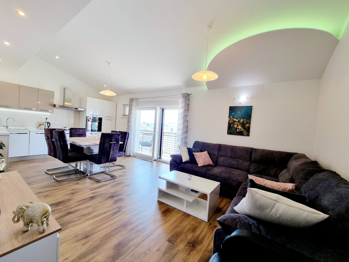 The spacious living area features modern furnishings, including a large sectional sofa and sleek dining chairs around a table. Natural light fills the space through glass doors leading to a balcony, while decorative accents create a homely feel.