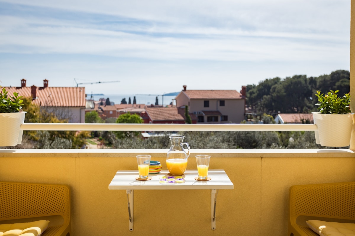A small terrace is depicted with a white table and two yellow chairs. On the table, a glass pitcher and two glasses of orange juice are set. The view showcases a peaceful landscape with houses and the sea visible in the distance.