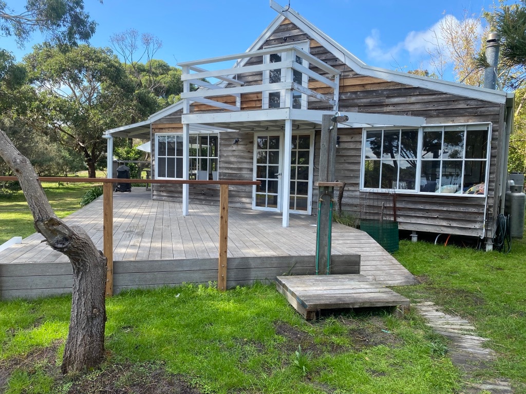 The cabin features a large outdoor deck area, which extends from the main entrance. The structure, made of timber, showcases multiple windows, allowing natural light to fill the space. Scenic greenery surrounds the cabin, enhancing the tranquil outdoor setting.