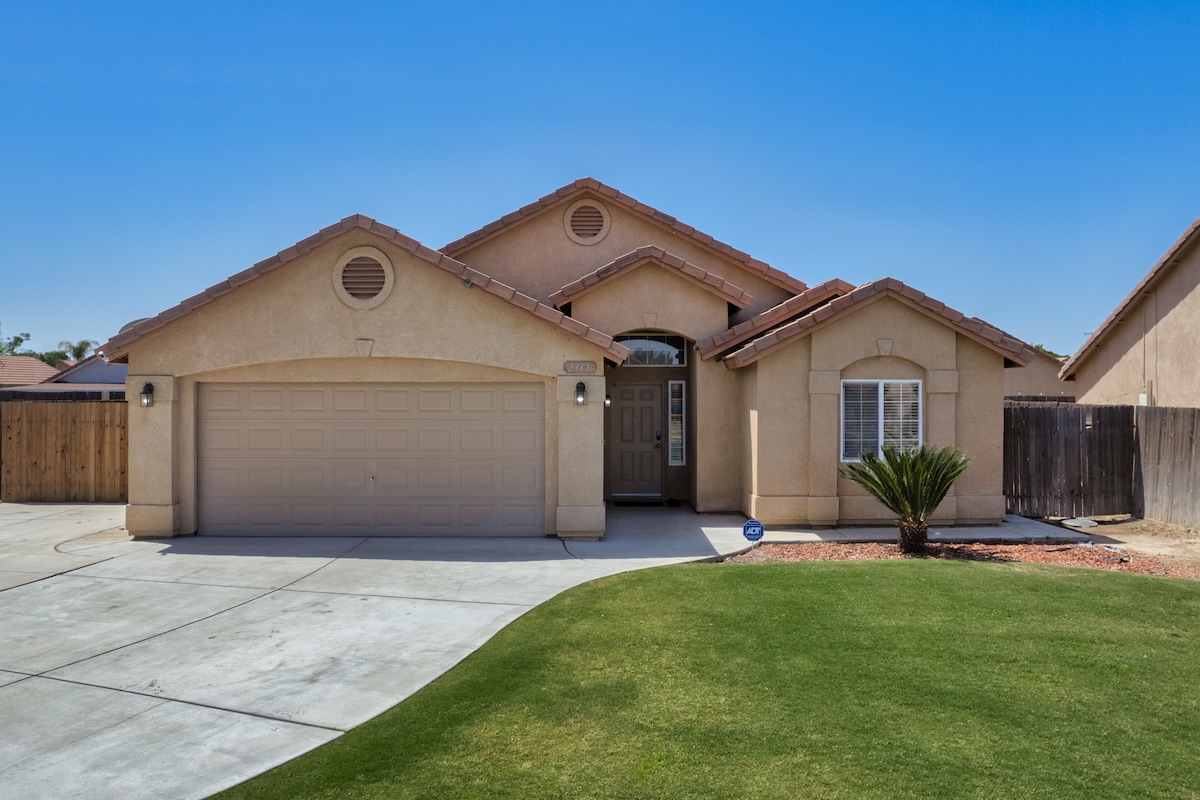 The exterior of the house features a single-story design with a double garage and a landscaped front yard. A pathway leads to the entrance, showcasing a warm color palette of neutral tones. Surrounding fences provide privacy, and a palm tree adds a touch of greenery.