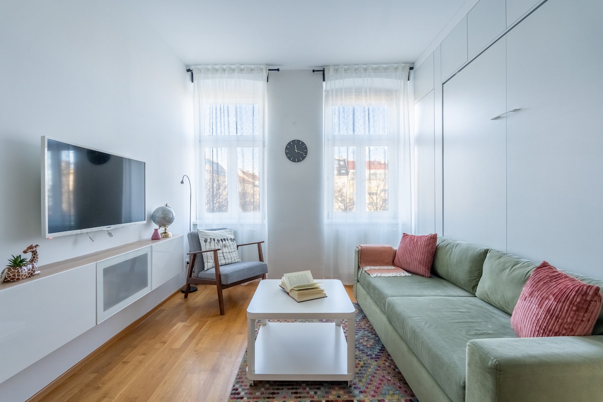 A bright living area features a comfortable green sofa and a light coffee table. Two large windows allow natural light to fill the space. A wall-mounted television is positioned above a sleek storage unit, and a small armchair adds an additional seating option.
