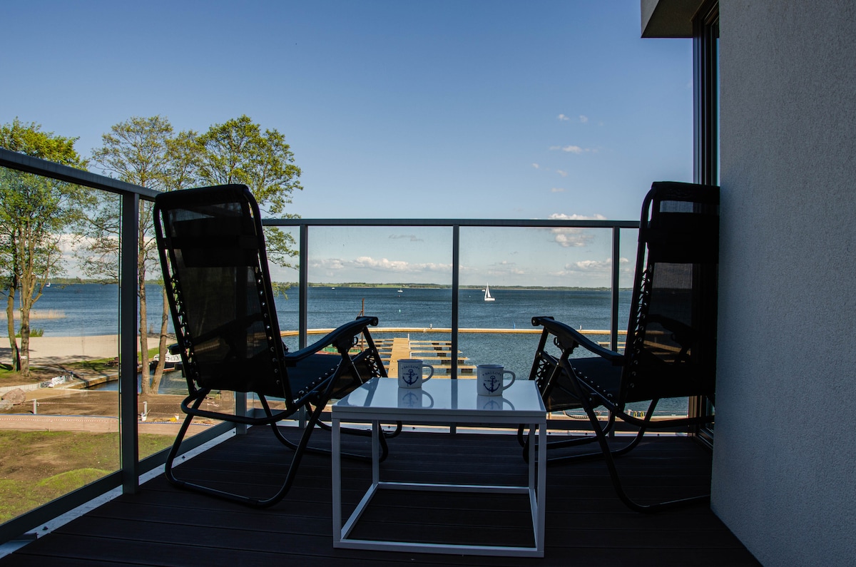 A serene balcony is presented with two comfortable chairs and a small table, overlooking a tranquil lake and marina. Clear skies and gentle waves are visible, creating a relaxed atmosphere for outdoor enjoyment.