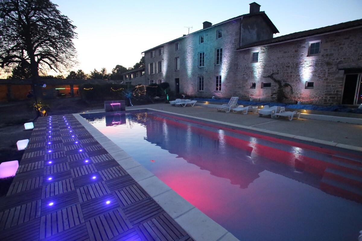 An inviting pool area is depicted, featuring a heated swimming pool surrounded by illuminated decking and lounge chairs. The ambiance is enhanced by colored lights reflecting off the water, while the historic stone building stands gracefully in the background under a dusky sky.
