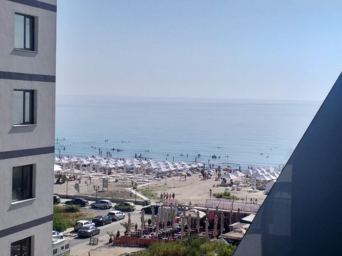 A clear view of the beach and shoreline is presented, showcasing calm water reflecting the sky. Several sunbeds and umbrellas are arranged along the sandy beach. People can be seen wading in the water, while beachside establishments are visible in the foreground.