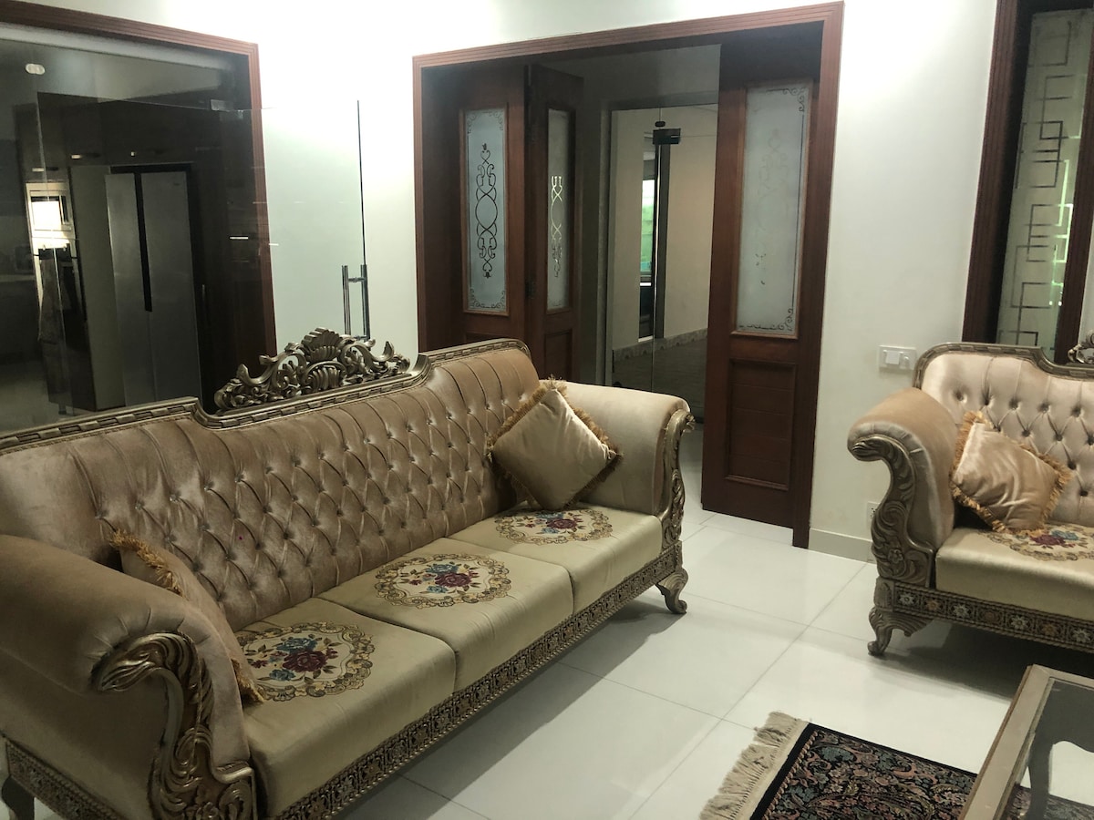 A spacious living area features elegant upholstered sofas with intricate detailing and decorative pillows. A mirrored wall reflects the soft lighting, enhancing the openness of the space. In the background, glimpses of a modern kitchen can be seen through double doors.