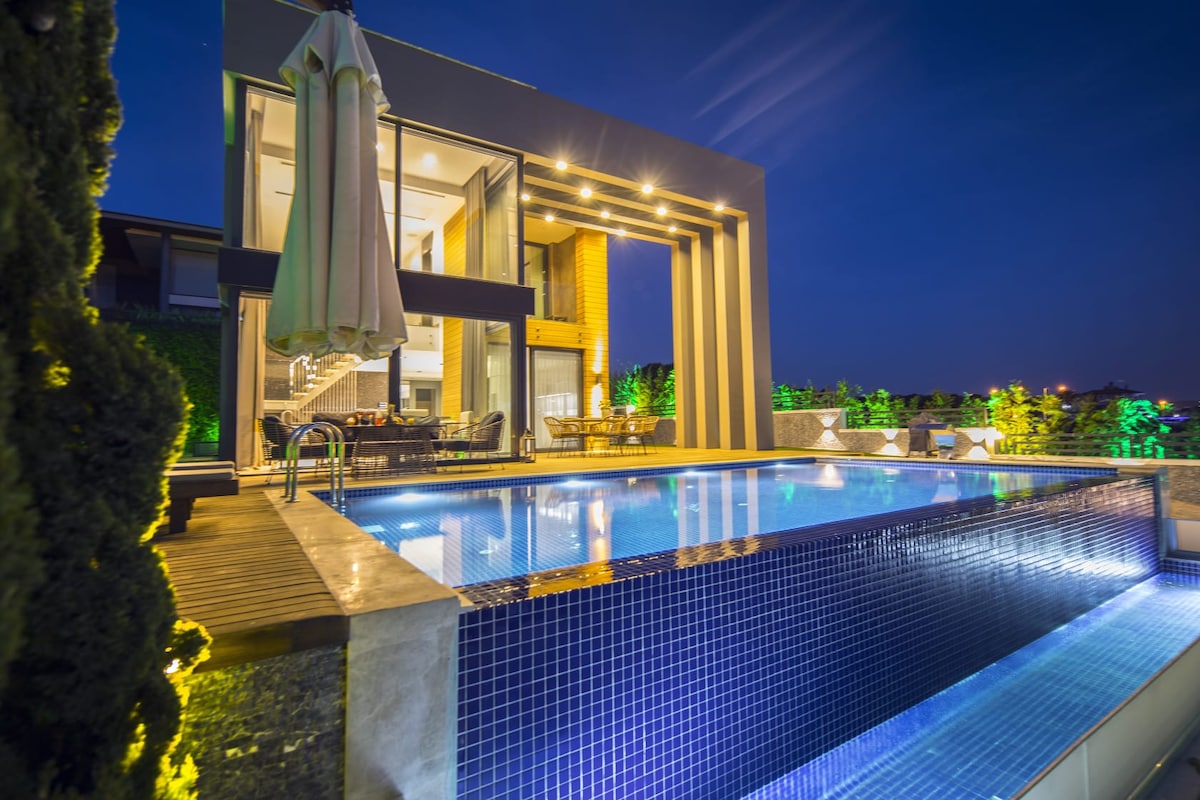 An outdoor terrace features a modern swimming pool, framed by stylish seating and illuminated by soft lighting. The architectural design includes large glass doors leading to the indoor living space, enhancing the connection between indoor and outdoor areas.