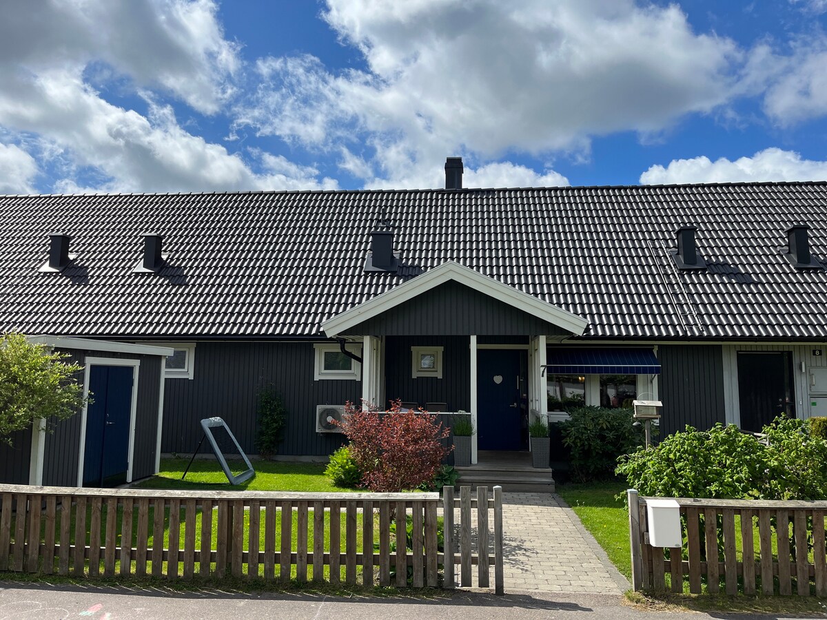 A two-story house features a dark exterior with a contrasting white trim. The sloped roof is covered with tiles, and a front porch leads to the entrance. Lush green grass surrounds the building, complemented by a small fence and a swing set in the yard.