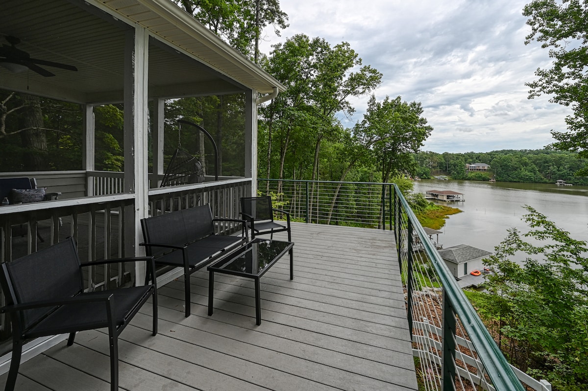 A spacious outdoor deck with contemporary seating arrangements, including black chairs and a glass table, provides a serene view of the lake and surrounding woods. A screened-in porch is visible, adding to the inviting outdoor space.