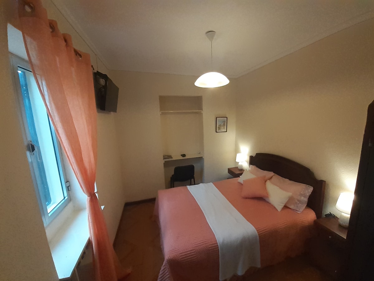A cozy bedroom features a spacious bed covered with a light-colored blanket and pillows. Natural light filters through a window adorned with orange curtains. A television is mounted on the wall, while a small desk and chair are present near the entrance.