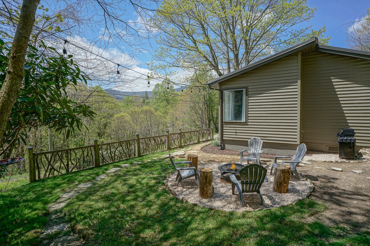 An outdoor gathering area features rustic seating arranged around a stone fire pit, surrounded by a grassy yard. String lights are draped overhead, enhancing the inviting atmosphere. A grill is positioned nearby, with scenic mountain views visible in the background.
