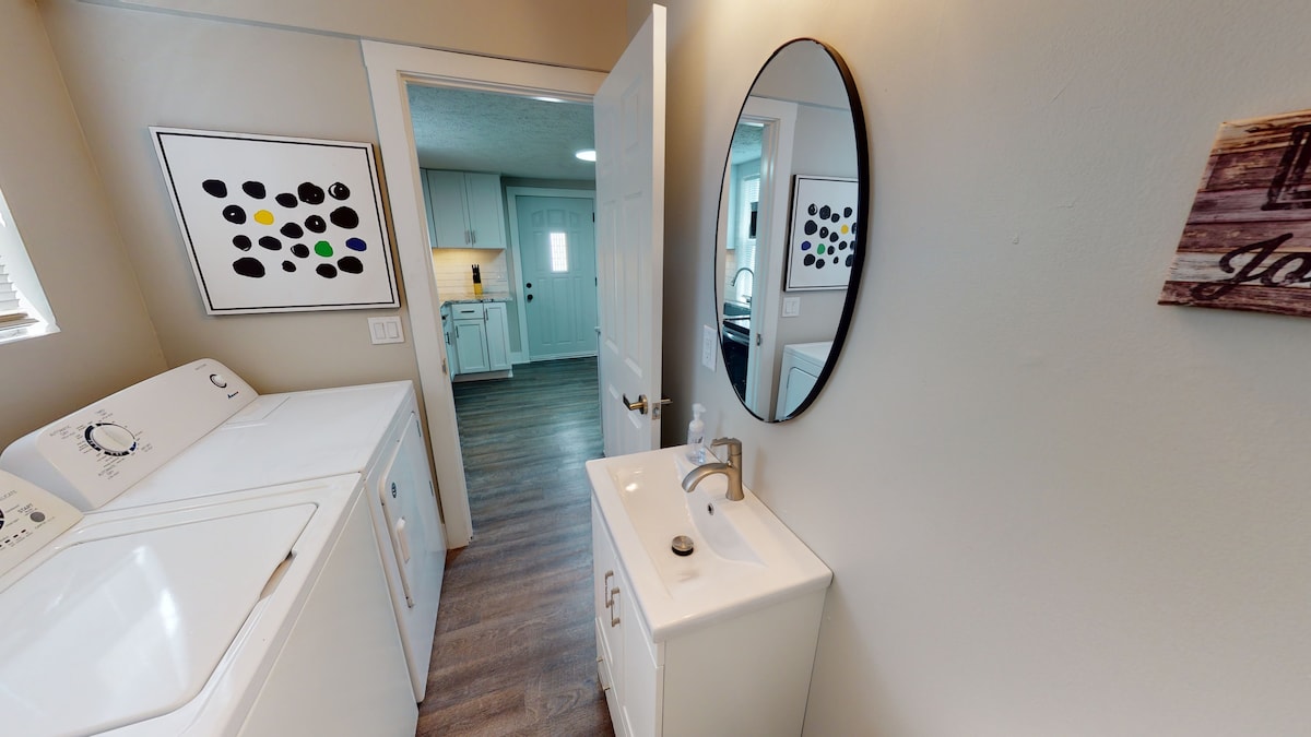 A functional laundry area is visible, featuring a stacked washer and dryer alongside a compact sink with a circular mirror above. Natural light enters through a nearby window, illuminating the clean lines and neutral tones of the space, leading into an adjoining kitchen area.
