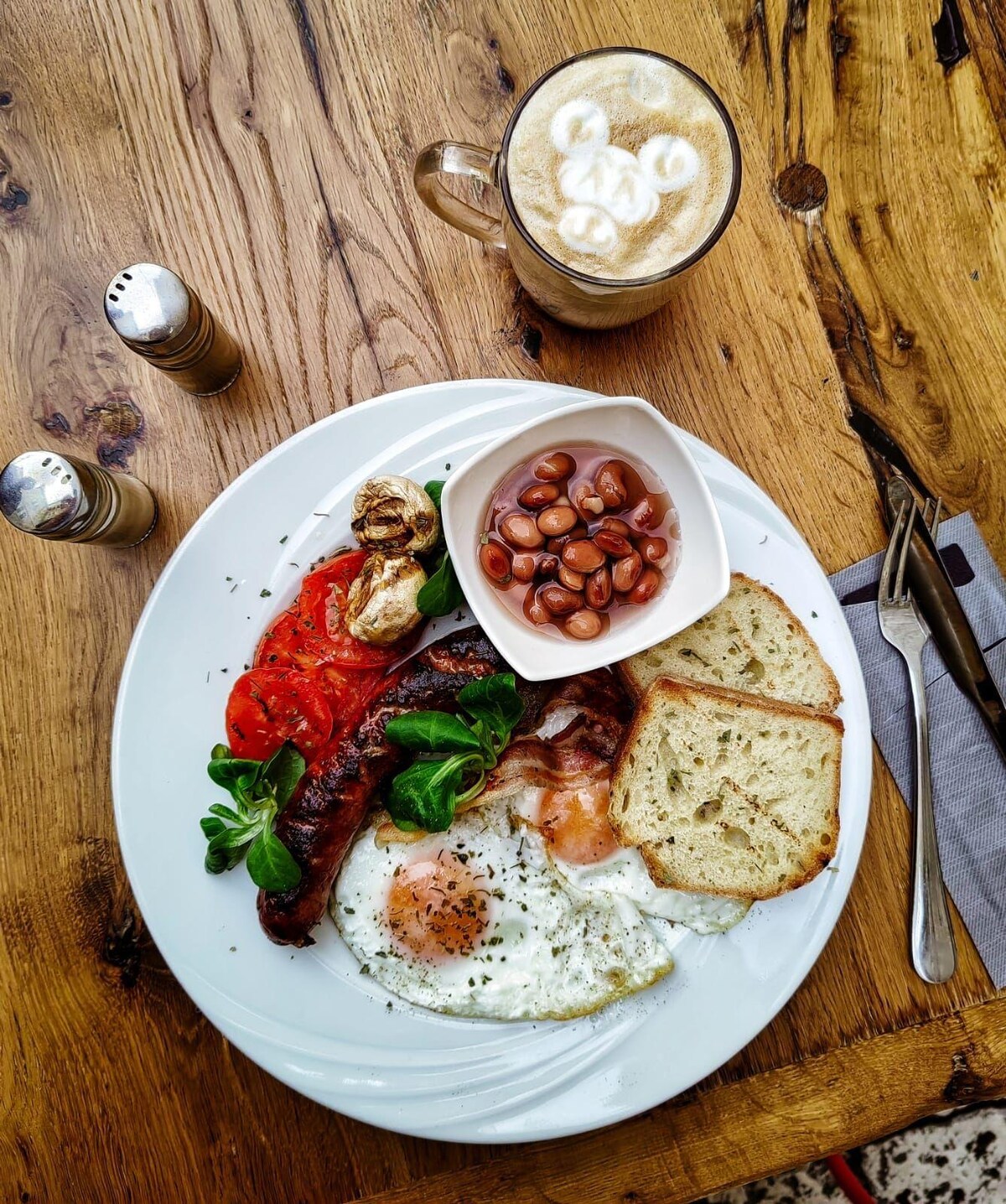 A plate of breakfast features two sunny-side-up eggs, grilled sausage, crispy bacon, sliced tomatoes, baked beans, and a slice of bread. The meal is presented on a round white plate, with a glass of coffee topped with foamed milk in the background.