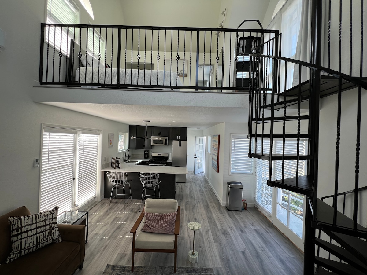 A spacious loft is shown with a spiral staircase leading to a sleeping area above. The open living and dining space features modern kitchen cabinets and seating for four. Ample natural light enters through large windows with blinds, enhancing the contemporary design.
