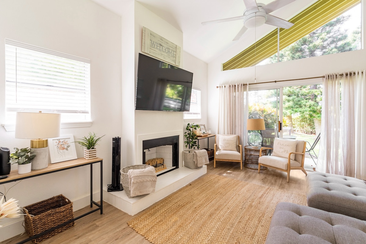 The living area is filled with natural light from large windows and a glass door leading to the outdoor space. A comfortable couch and armchair are arranged around a low coffee table. A mounted TV is situated above a modern fireplace. A jute rug adds texture underfoot.
