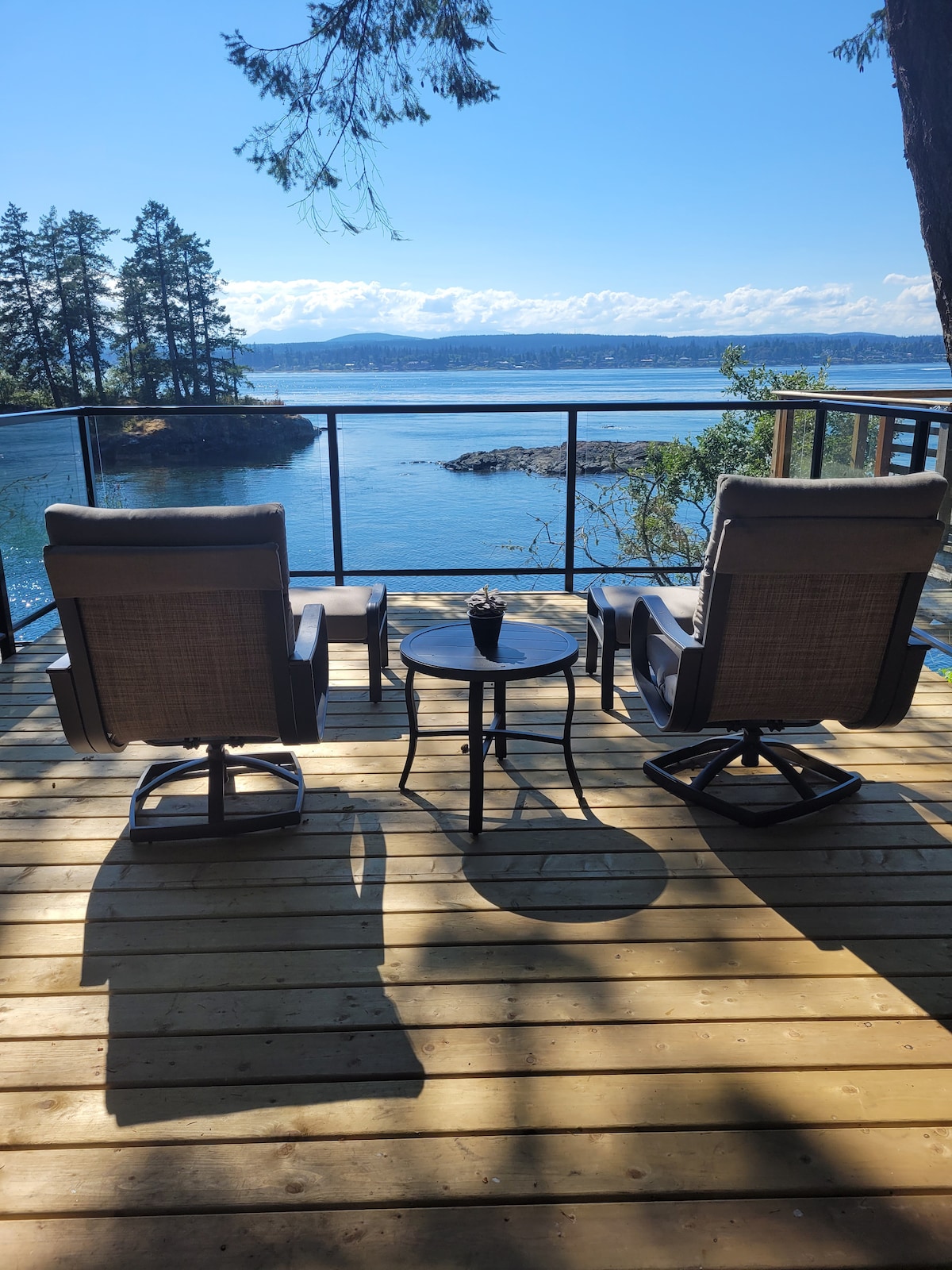 Two comfortable chairs are positioned on a wooden deck overlooking the ocean, accompanied by a small round table. The clear blue water reflects the sky, and trees frame the view, providing a serene outdoor space for relaxation.