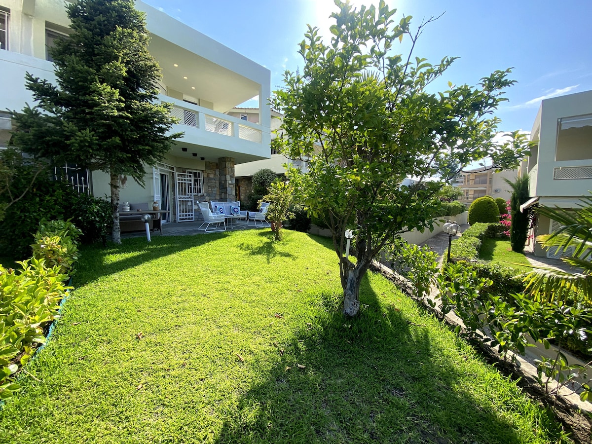 A lush green garden area is highlighted, featuring well-maintained grass and a small tree. Comfortable seating with sun chairs is positioned in the background, surrounded by an inviting environment, providing ample space for outdoor relaxation.