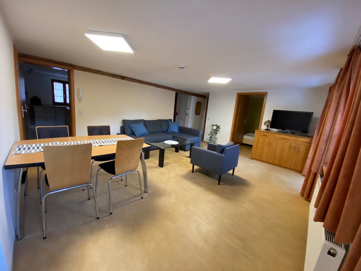 The living area is arranged with a comfortable seating arrangement featuring a dark blue sofa and two accent chairs. A wooden dining table with chairs is centrally placed, while a television stands against the wall beside a wooden cabinet. Natural light enters through a nearby window.