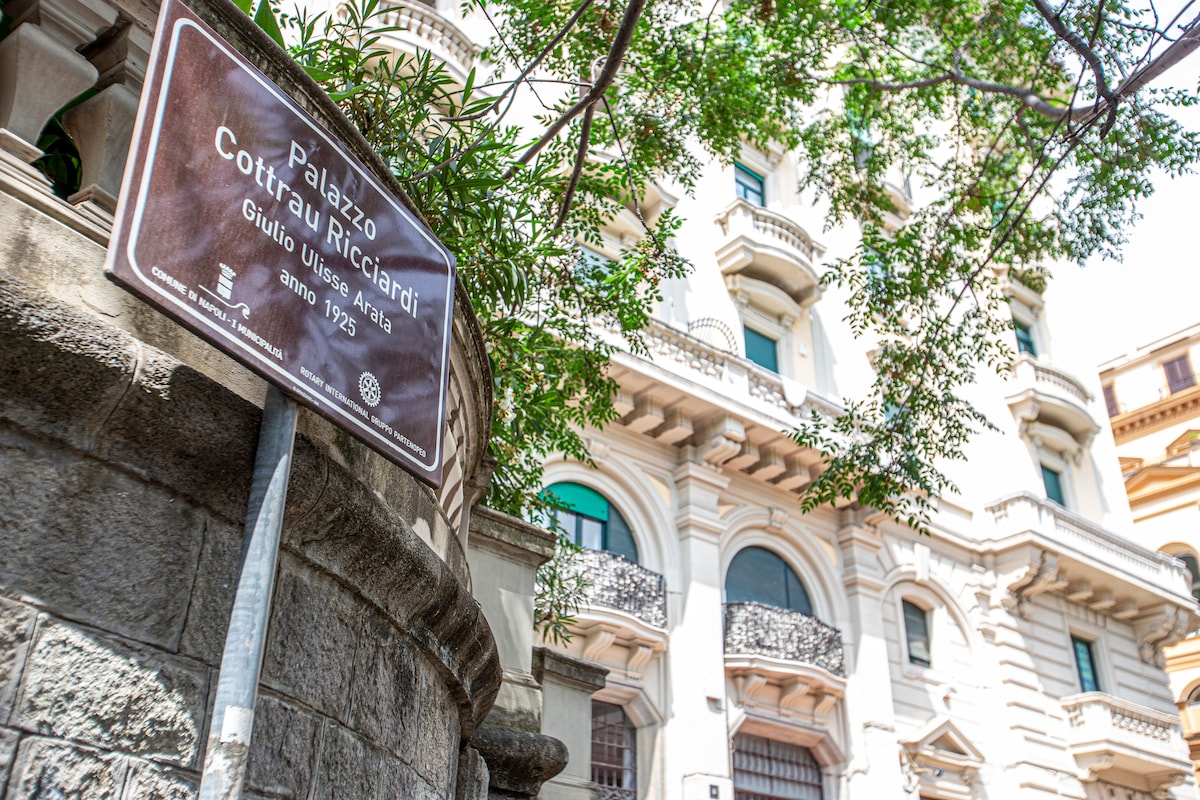 A historical building is showcased, reflecting architectural elegance with its ornate balcony and intricate stonework. A sign indicating 'Palazzo Cottura Ricciardi' is visible in the foreground, partially obscured by lush green leaves, emphasizing the building's grandeur.