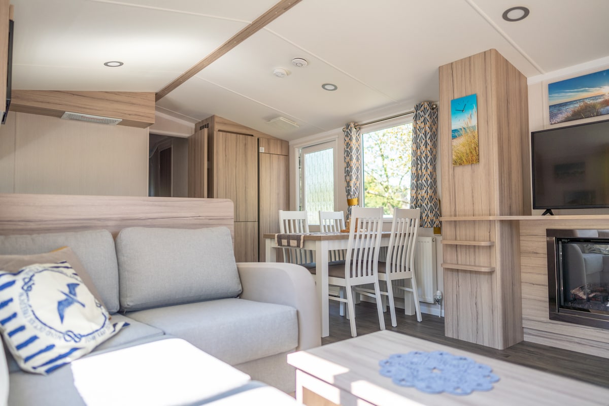 An inviting living area features a light-colored couch with decorative pillows, complemented by a wooden dining table surrounded by white chairs. Sunlight streams through windows adorned with patterned curtains, creating a cozy ambiance. A television and a decorative fireplace enhance the space.