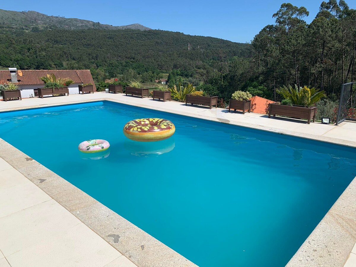 A private swimming pool is surrounded by a spacious terrace, featuring comfortable seating options and lush greenery. Colorful inflatable floats rest on the water's surface, against a backdrop of rolling hills and clear blue skies.