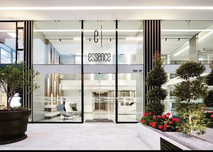 The building's entrance features large glass doors with the name 'el essence' prominently displayed. Surrounding greenery and decorative plants enhance the modern design, creating a welcoming entryway. The interior is visible through the glass, showcasing sleek furnishings and an open, airy atmosphere.