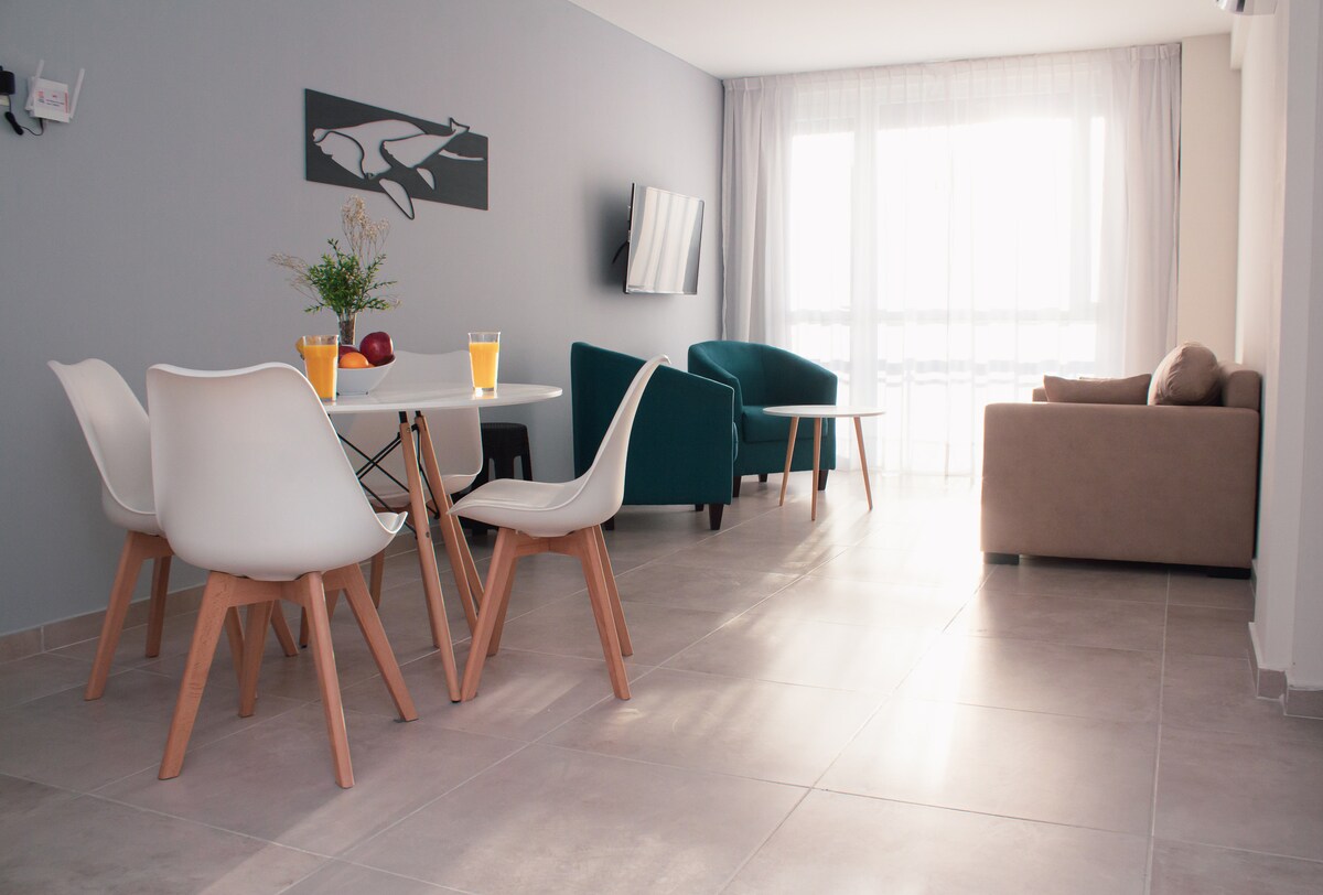 An open living space includes a dining area with a round table and six chairs. Two teal armchairs and a beige couch offer seating options. A television is mounted on the wall, while natural light enters through large sliding glass doors, enhancing the light-colored tile floor.