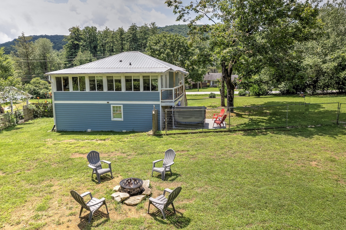 A fenced-in backyard features a fire pit surrounded by six Adirondack chairs on a grassy area. The cottage stands in the background, with a porch and large windows, framed by trees and mountains.