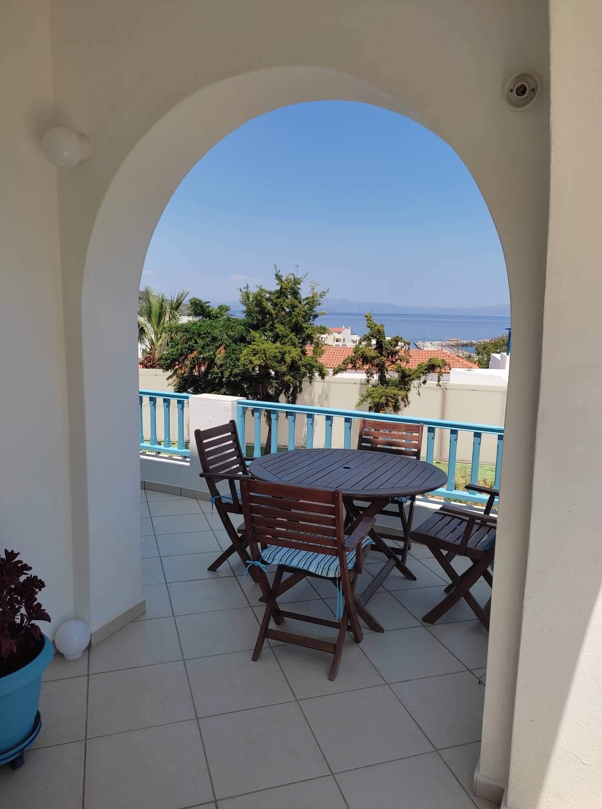 A balcony area is framed by an arched opening, featuring a round wooden table surrounded by four folding chairs. Potted plants add greenery, with a view of the sea visible in the background under a clear blue sky.