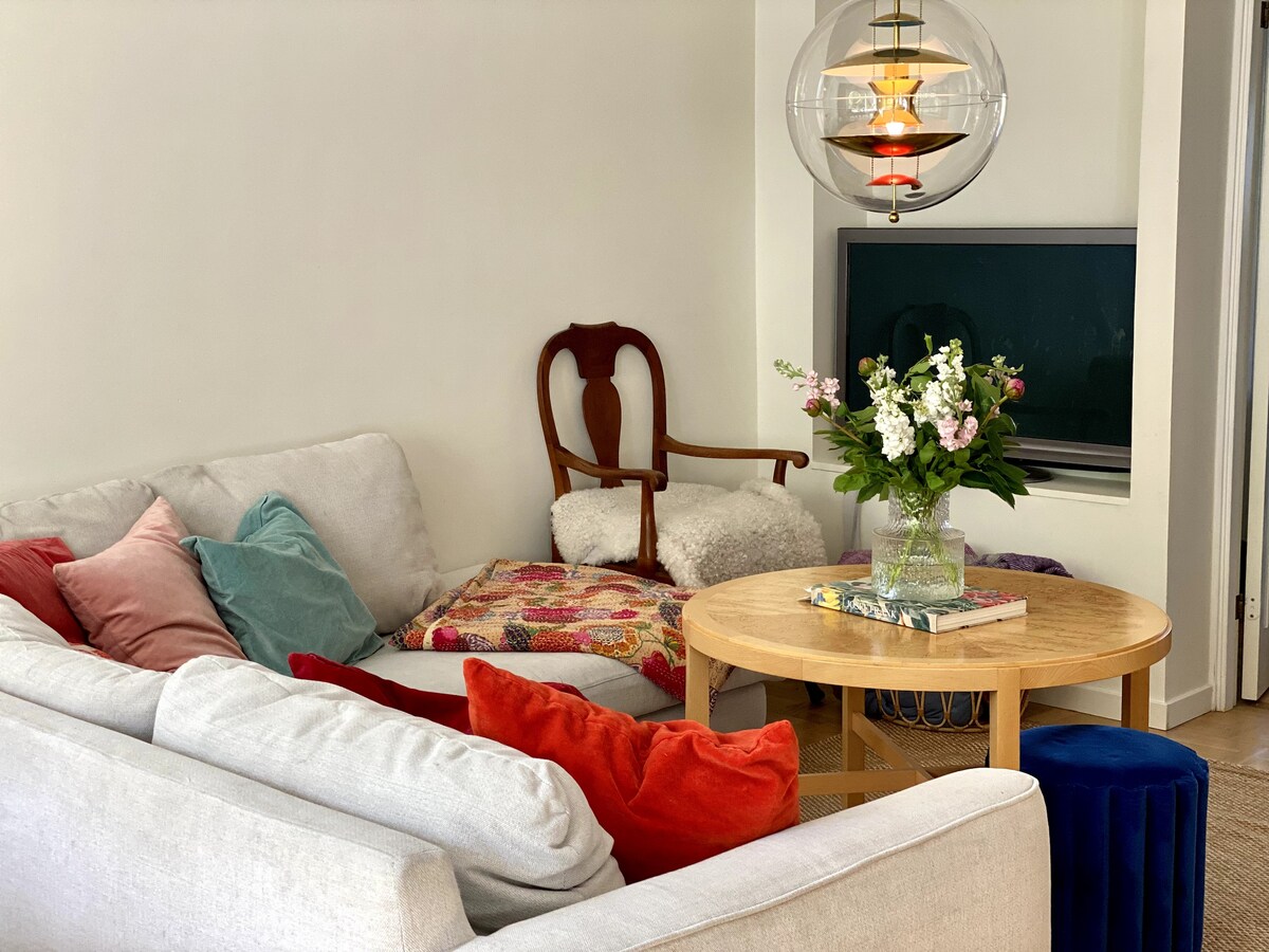 A comfortable seating area is presented with a light-colored sofa adorned with colorful pillows. A round wooden table is displayed centrally, complemented by a small, decorative floral arrangement. An armchair sits in the corner, while a modern light fixture hangs above the space.