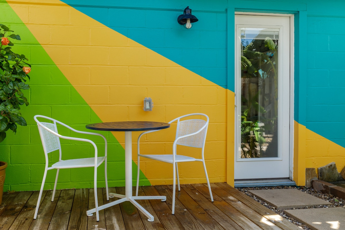 A cheerful outdoor space is featured, showcasing a round table surrounded by two white chairs. The vibrant wall in the background displays a bold design with shades of turquoise, yellow, and green, complementing the wooden deck below.