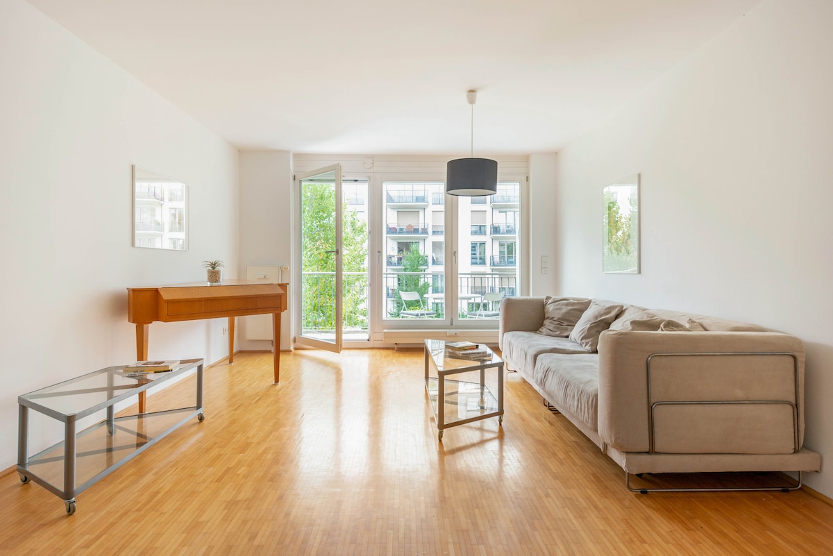 The living room features a light wooden floor and a spacious sofa. A glass coffee table and a wooden piano add to the simple decor. Large windows allow natural light to fill the room, with views of greenery from the balcony.