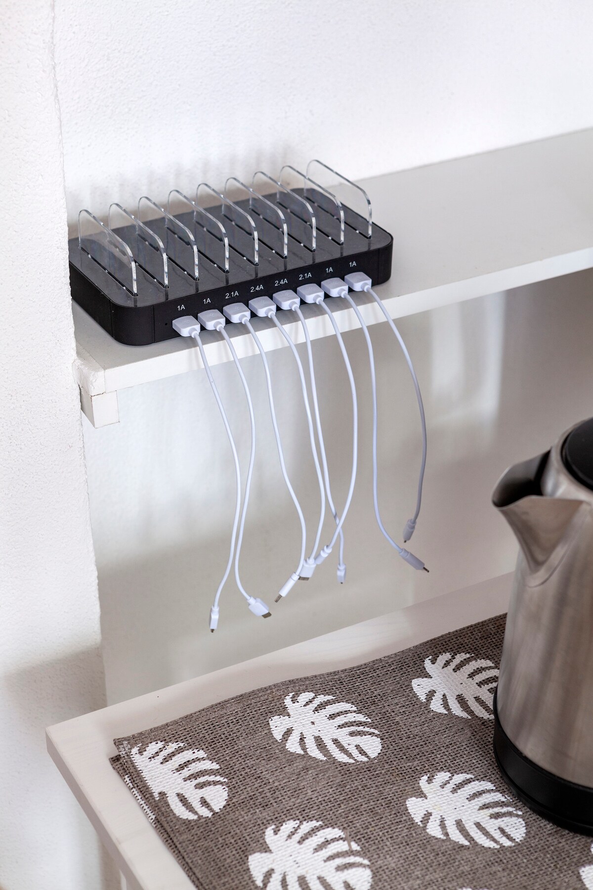 A charging station is positioned on a white shelf, equipped with multiple outlets and charging cables. A kettle is visible nearby, resting on a neutral-colored placemat featuring a leaf pattern. The setup supports easy access to power for devices.