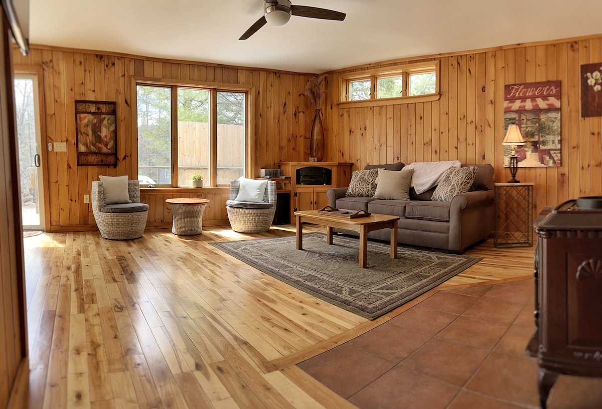 A spacious living area features wooden walls and flooring, creating a natural atmosphere. Two comfortable chairs are placed beside a table, while a sofa is positioned in front of large windows that offer outdoor views. A decorative rug lays on the floor, adding texture to the space.