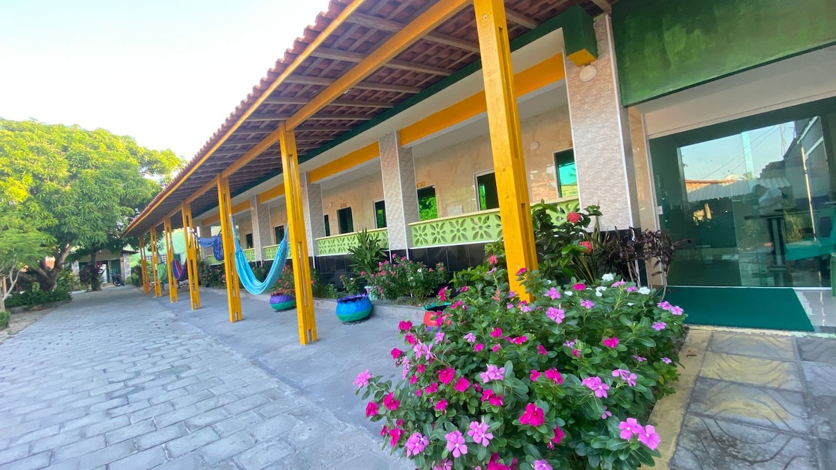 A pathway lined with colorful flowers leads to the entrance of a building featuring a vibrant facade. The yellow columns support a covered porch, with hammocks spaced along the walkway, creating a relaxed and inviting outdoor area shaded by trees.
