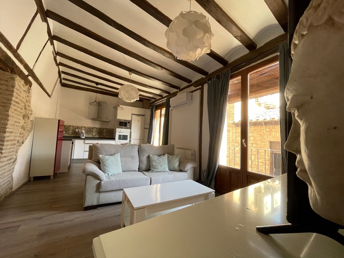 A cozy living area is highlighted with wooden beams and natural light from large windows. A soft sofa is positioned near a white coffee table. The kitchen is accessible, showcasing modern appliances and stylish cabinetry. Decorative light fixtures add a contemporary touch.