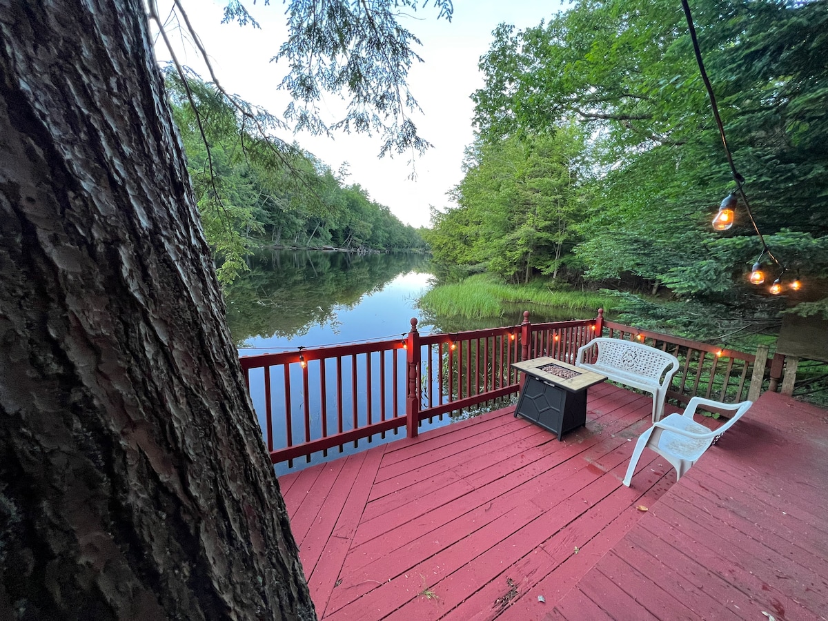 A red deck offers serene water views, surrounded by green foliage. A small table and a white chair are positioned on the deck, creating a peaceful outdoor space. Soft lighting from string lights adds a warm touch to the setting.