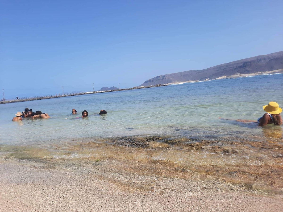 A sandy beach area is visible, with a clear body of water gently lapping at the shore. Several people can be seen enjoying the water, with some standing and others swimming. In the background, a rocky landscape and a distant pier are noted under a clear blue sky.