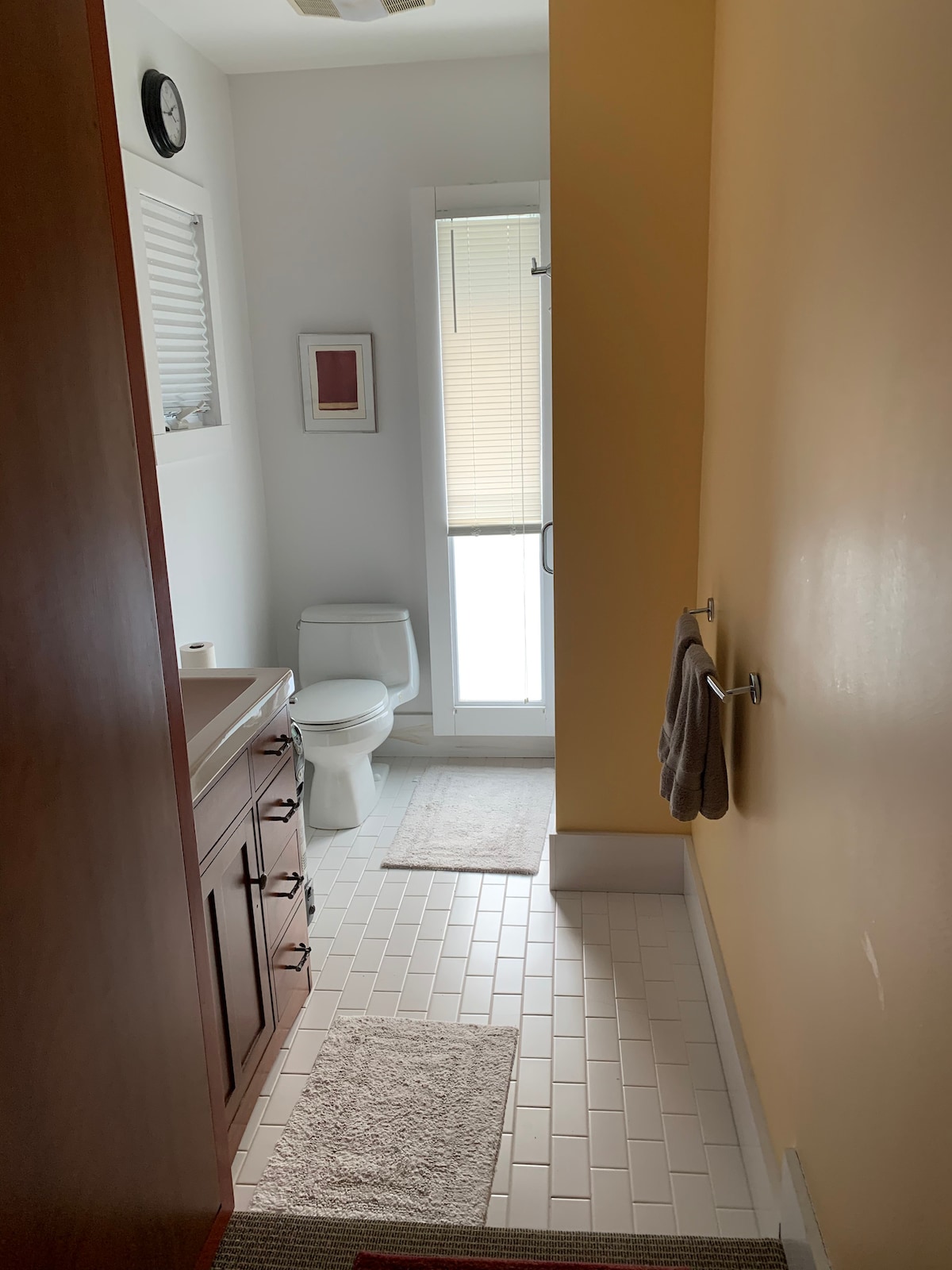 A bright and modern bathroom is shown, featuring a white tiled floor and warm yellow walls. A large mirror is mounted above a wooden vanity with ample storage. Soft towels hang on a polished fixtures, and natural light filters through a partially obscured window.