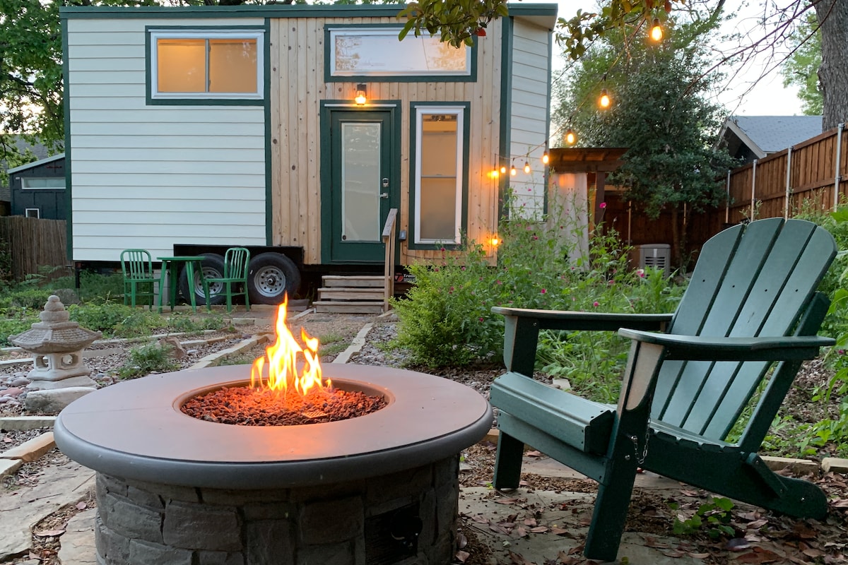A cozy outdoor firepit surrounded by comfortable green seating is visible in the foreground. The well-lit tiny house stands in the background, showcasing its light green exterior and large windows. Lush greenery and gentle landscaping create a welcoming atmosphere.
