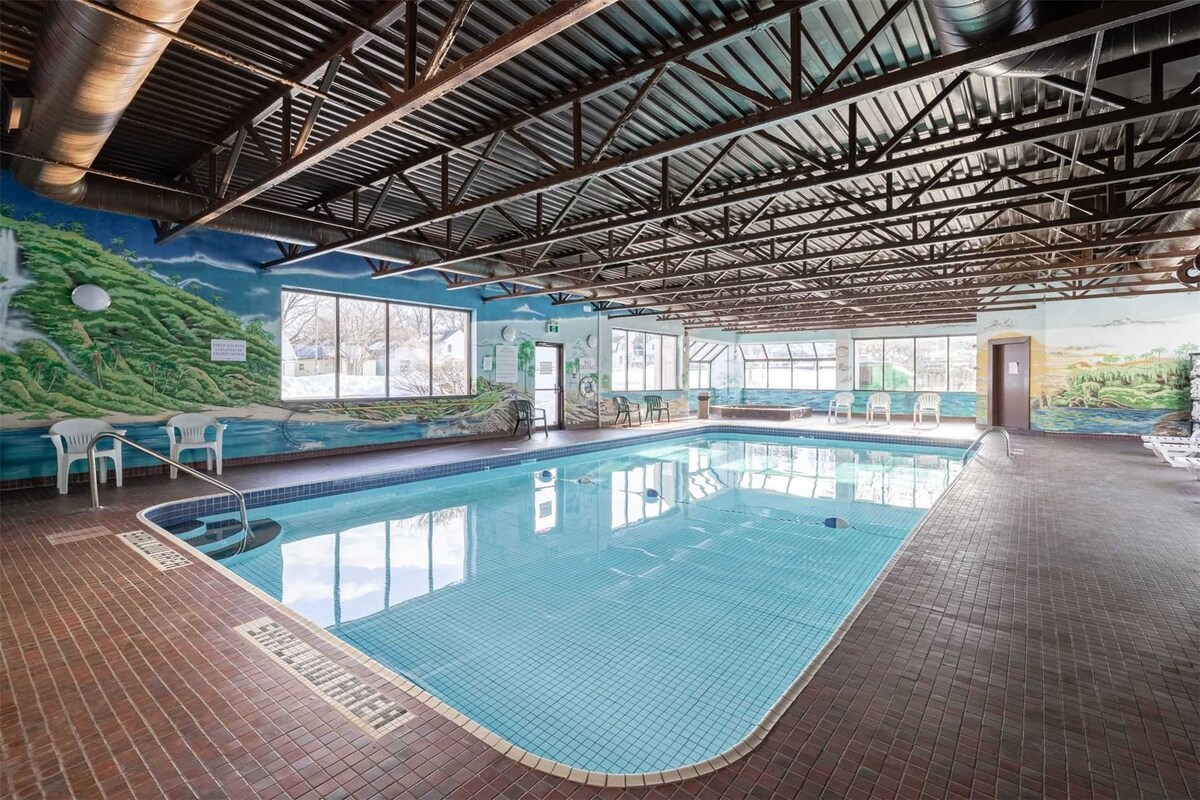 An indoor swimming pool is surrounded by large windows, providing ample natural light. The pool area features a mural depicting natural scenery. Several white chairs are arranged around the pool, and the floor is covered in dark tiled flooring.