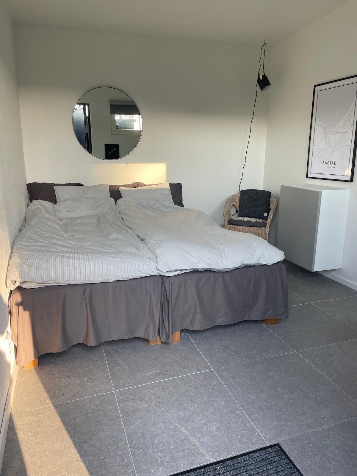 A spacious bed with light-colored bedding is positioned in a room featuring soft natural light. A round mirror hangs above, while a cozy chair and a minimalist cabinet complete the simple decor, all set against neutral walls and a sleek tile floor.