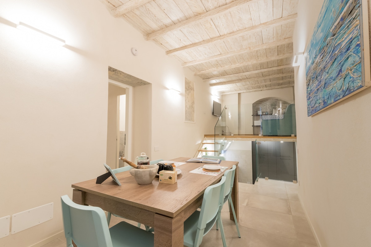 The spacious dining area features a wooden table surrounded by light blue chairs. A modern flat-screen TV is mounted on the wall, and a staircase leading to the upper level is visible in the background. Soft lighting and neutral tones create an inviting ambiance.
