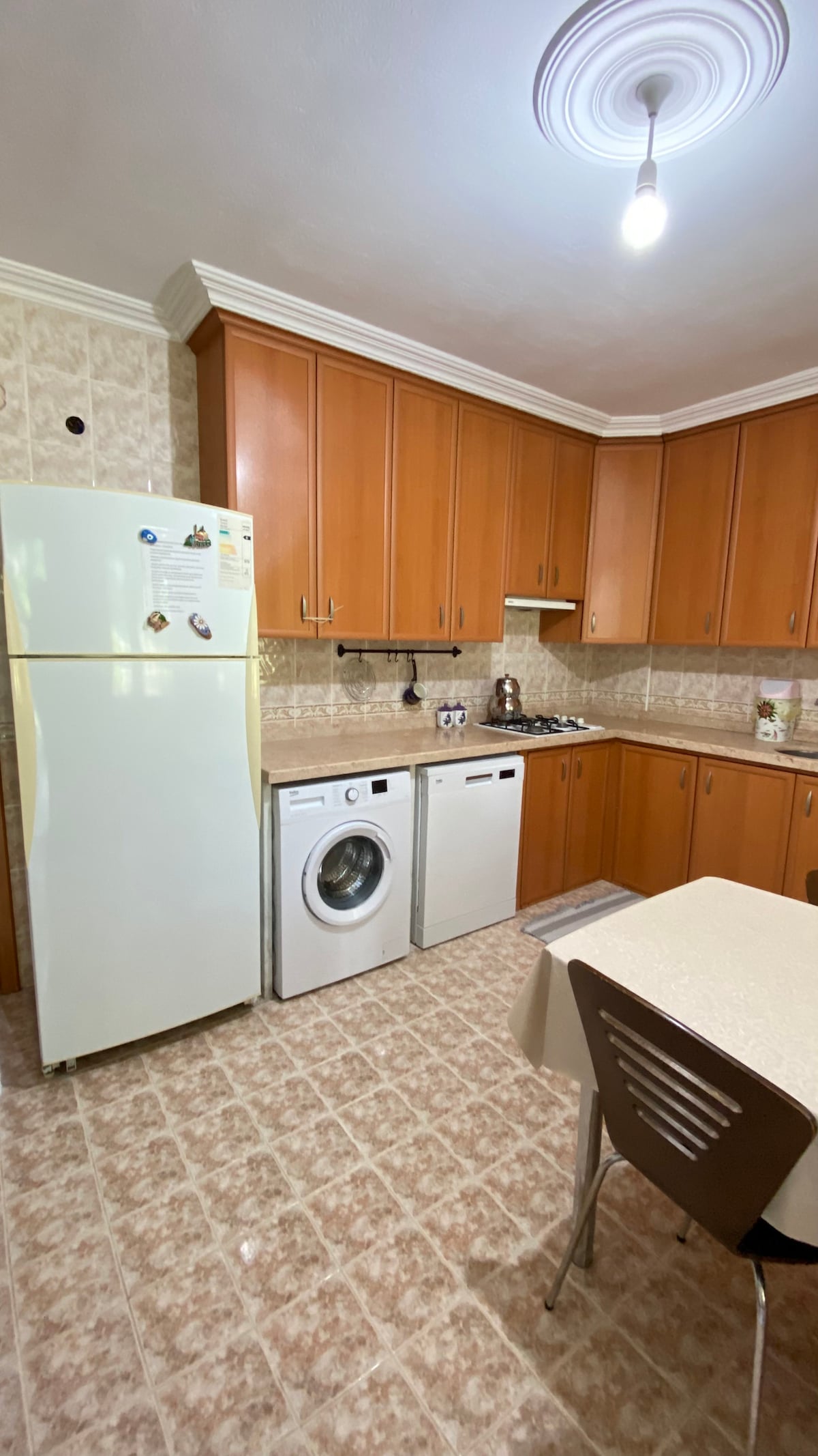 The kitchen is equipped with wooden cabinets and a white refrigerator. A washing machine and a dishwasher are positioned next to the sink. A small dining table with two chairs is situated near the wall, creating a functional and organized space.