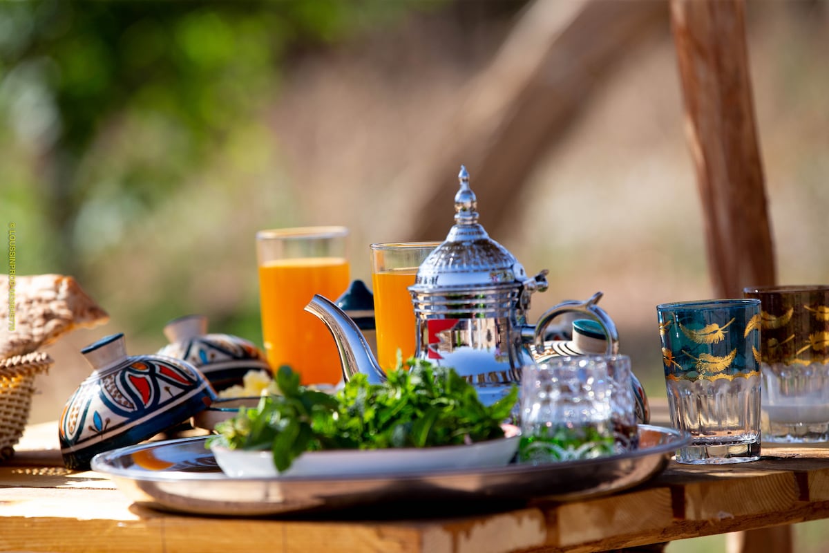 An outdoor table is set with a tray featuring ornate pottery and a traditional teapot. Glasses filled with orange juice reflect sunlight, and fresh herbs are arranged on the platter beside woven baskets, creating a charming and inviting dining display.