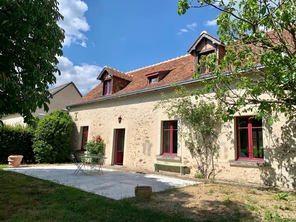 The exterior of the house is displayed, featuring a stone façade with large windows framed in red. A small seating area with two chairs and a table is visible on a patio under a blue sky, surrounded by lush green shrubs and trees.